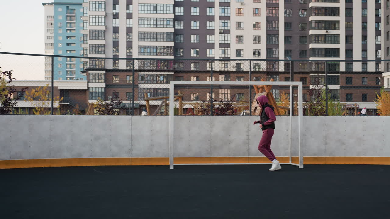 Female jogger wearing hoodie vest and joggers sneakers runs around circular outdoor sports court marked by white post under modern apartment building facades at dusk in urban setting