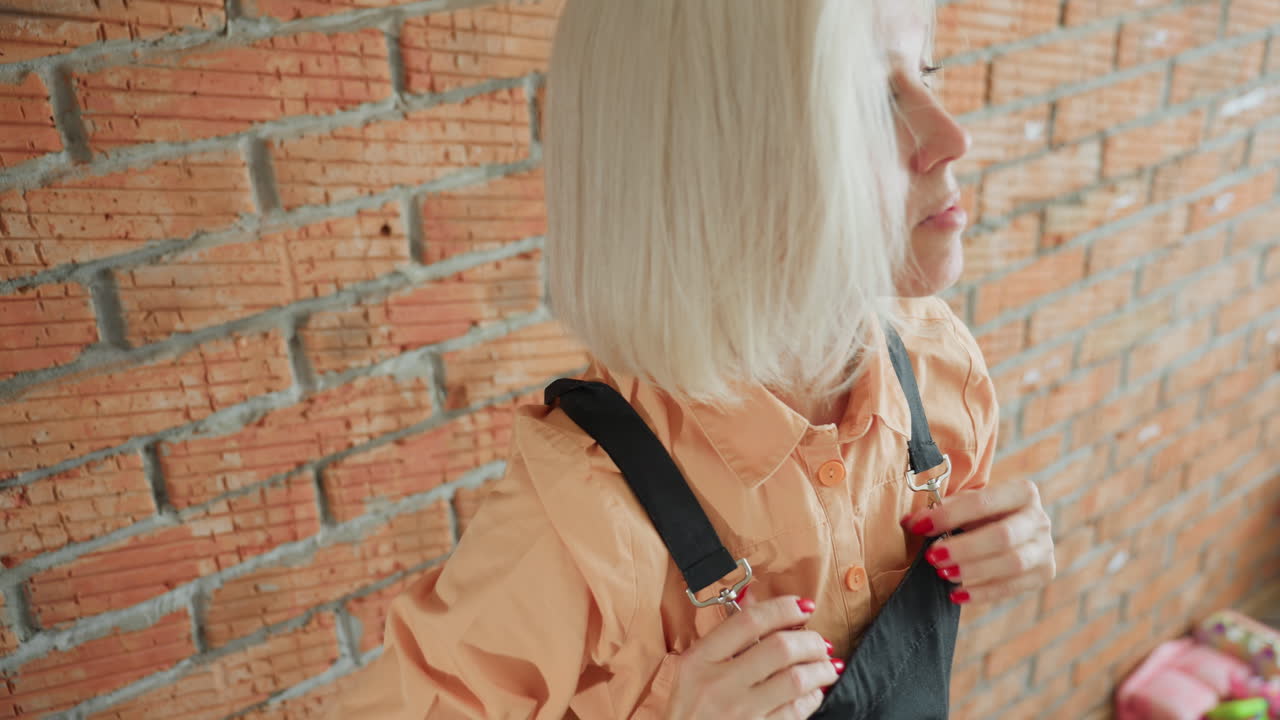 Decorator woman adjusting black apron over orange shirt standing against rustic brick wall, preparing for creative work session with focused expression, readiness, and artistic dedication