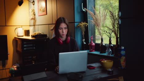 una chica morena con auriculares rojos trabaja en una computadora portátil en una habitación oscura con una ventana con vistas a un bosque verde