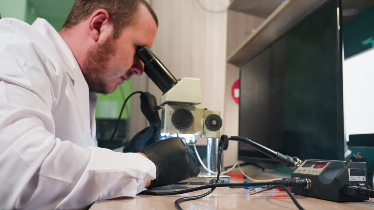 Close view of a technician in a white lab coat using a microscope having light to work on circuit board, the workbench is filled with various tools