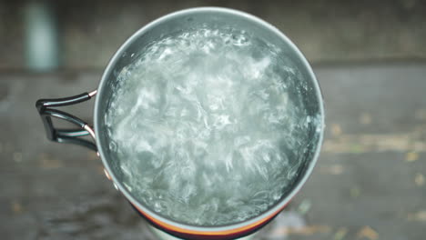 Close-up-overview-of-travel-gas-burner-and-small-pot-with-boiling-and-steaming-hot-water-at-Glacier-NP-Campground-and-Creek-camp-site-in-USA