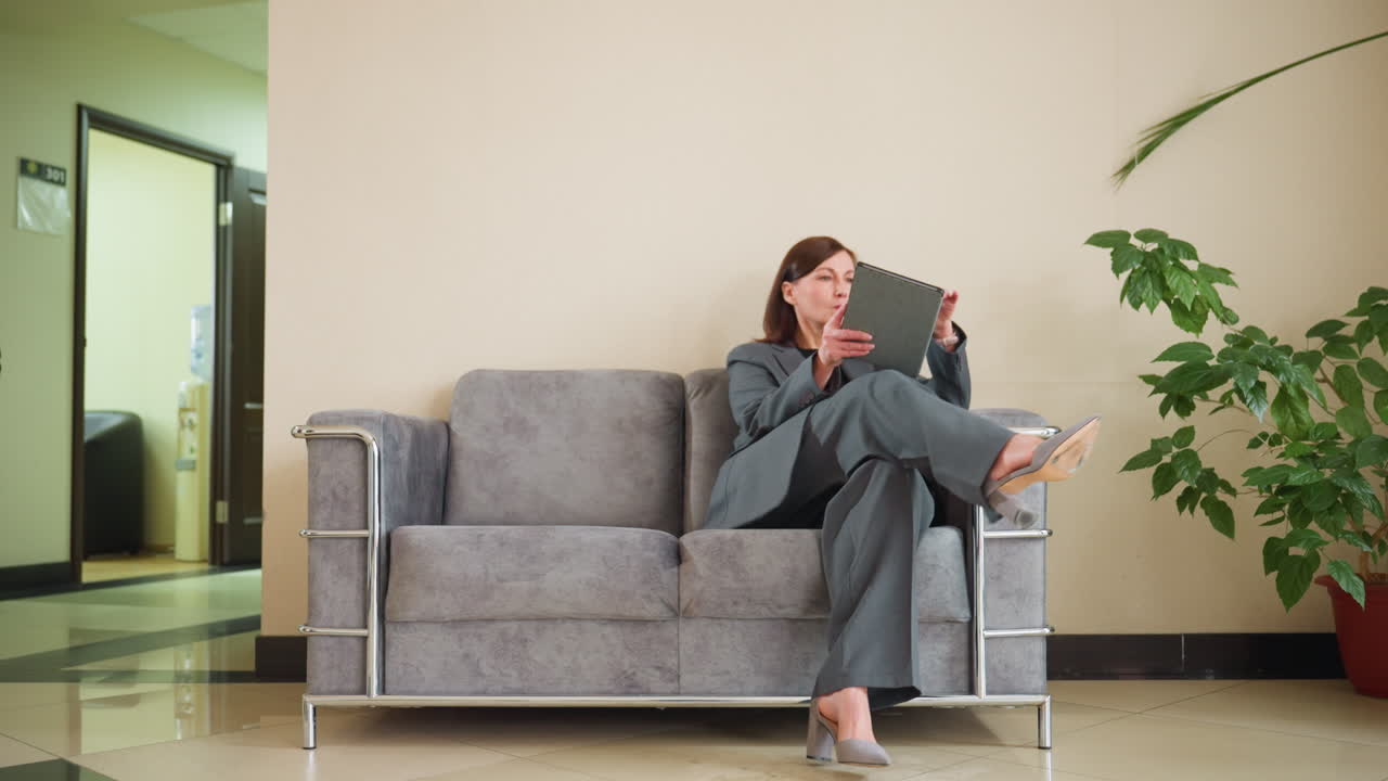 Business professional in gray suit walking through modern office lobby holding notebook. Person passing stylish minimalist furniture, standing near contemporary couch with metal armrests
