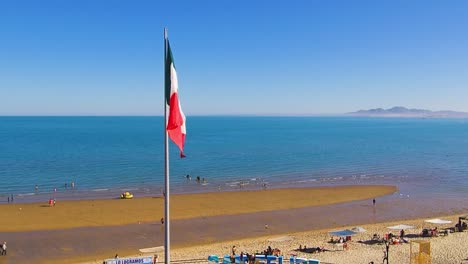 Vista-De-Un-Dron-Volando-A-La-Derecha-Mostrando-Una-Playa-Y-La-Bandera-Mexicana