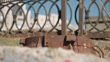 close-up view of a rusty metal object on the ground with an ornate fence and urban buildings in the background, with a passing by