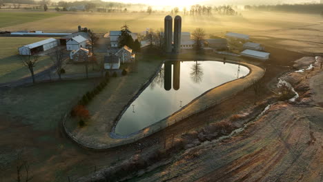 Establishing-shot-of-farm-with-silo-reflections-in-pond-water-at-winter-sunrise