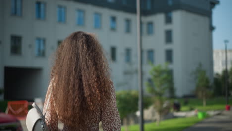 back view of young woman in vintage clothing walking outdoors, adjusting her curly hair, the scene features a blurred background with modern buildings, lush greenery, and a light pole