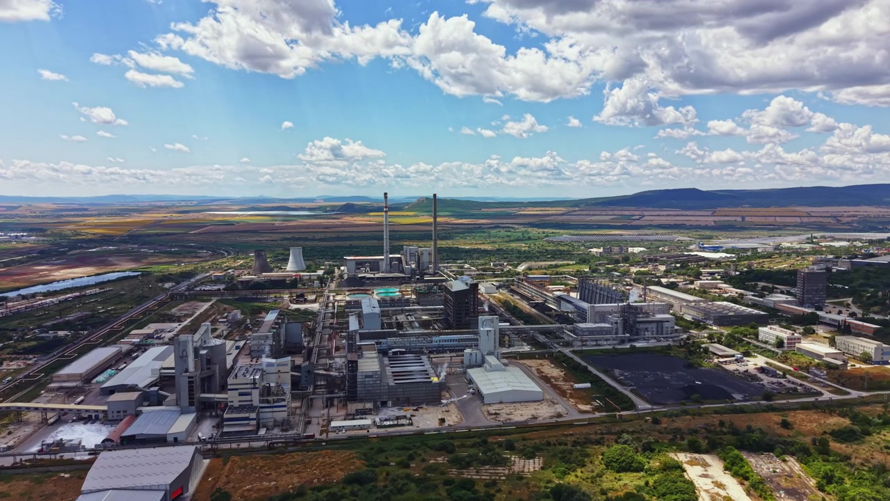 Vista di un grande complesso industriale situato in un'area rurale con edifici, macchinari e terreno aperto sotto un cielo nuvoloso vicino alle montagne.