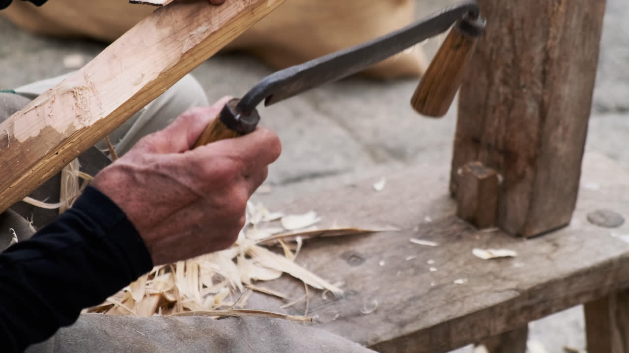 Bottom-to-top pan following an elderly artisan shaving a log with a drawknife to create wood splints for basket weaving; curling chips, textured hands, outdoor daylight