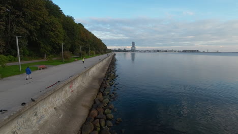 People-walking-on-Boulevard-in-Gdynia,-low-drone-flight-over-coast,-birds-start-from-off-the-coast