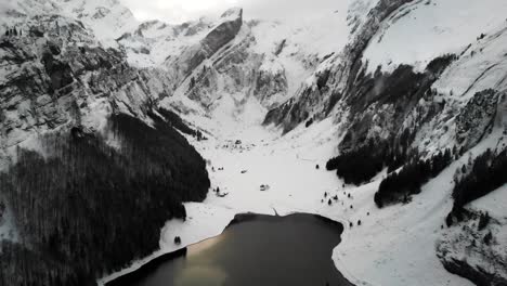 Aerial-flyover-over-lake-Seealpsee-in-Appenzell,-Switzerland-with-a-winter-landscape-full-of-snow-and-views-of-cliffs-and-peaks-of-Alpstein