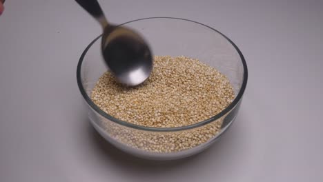 a small clear glass bowl filled with raw uncooked white quinoa. a hand holding a stainless steel spoon comes into frame and starts moving the quinoa grains around with the spoon