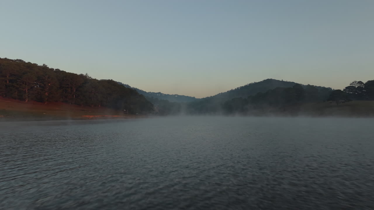 Early morning mist floats over a calm lake between forested hills