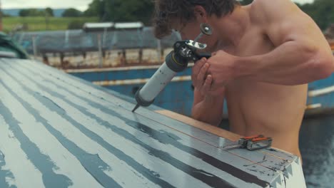 topless young man using manual sealant gun to seal cabin roof planks on wooden boat, mid shot