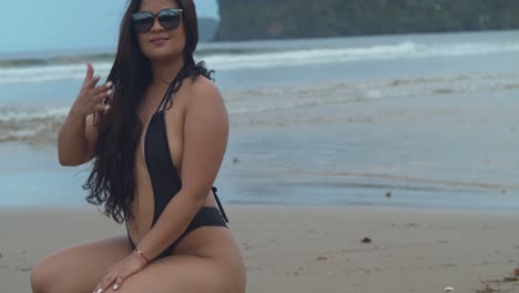 hispanic bikini babe sits at the shoreline and plays in her hair while watching the waves crash at the shoreline