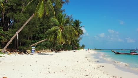 Paradise-tropical-island-with-white-sandy-exotic-beach-under-palm-trees-with-coconut-seed-hanging-over-turquoise-lagoon-with-boats,-Thailand