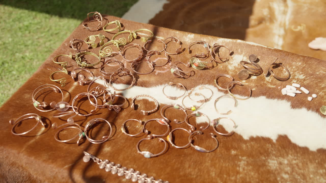 A slow dolly shot moves over a display of handmade historical replica jewelry. The collection of bronze and copper rings and bracelets is laid out on an animal hide