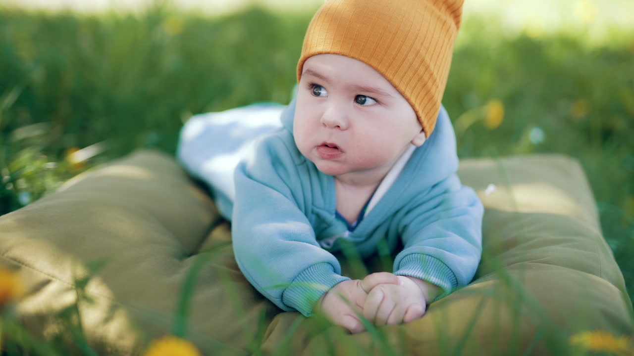 Little baby boy in blue clothes and orange cap lies on the pillow in green grass. Sweet kid yawns and looks around. Blurred backdrop.
