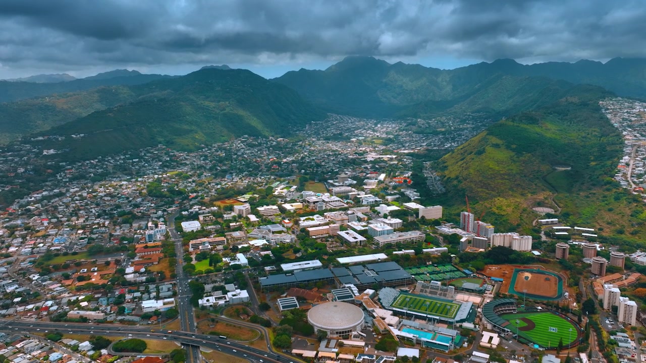 Nuvole piovose si addensano sopra Honolulu, circondata da montagne verdeggianti. Riprese aeree in avvicinamento allo stadio dell'Università delle Hawaii. Veduta aerea di Honolulu