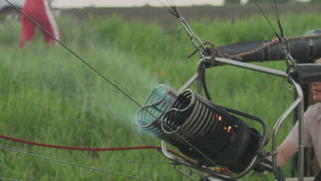 young man lifts metal burner while intense flame bursts from hot air balloon engine during setup on grassy field, showing strong fire pressure, fuel ignition, and balloon inflation process in action