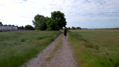 Fast-take-of-and-aerial-following-of-male-trail-runner-along-river-IJssel-in-Dutch-with-countenance-of-tower-town-Zutphen-behind