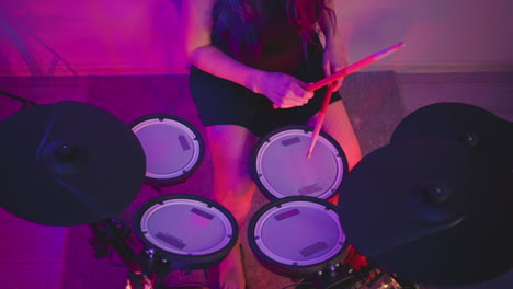 top down view of girl seated at electronic drum kit, holding sticks while practicing rhythm on drum pads, surrounded by pink and purple lighting in creative indoor studio environment