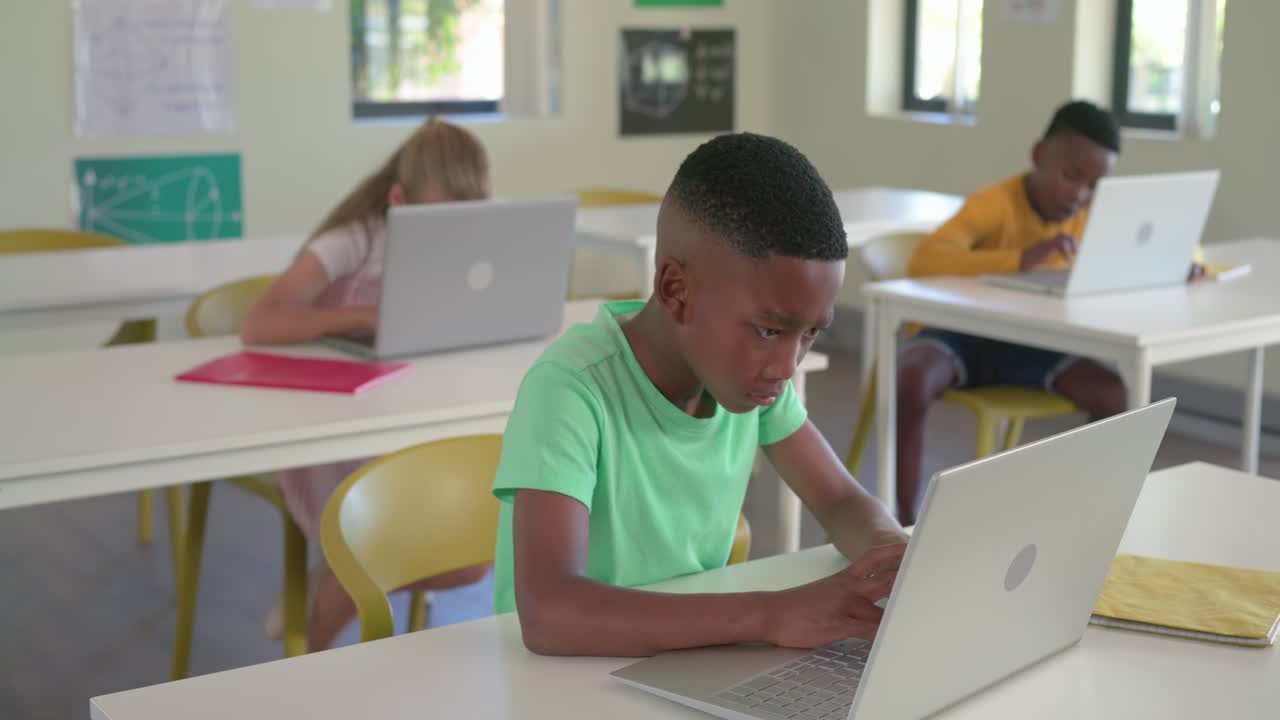 Ragazzo afroamericano con una maglietta verde che digita su un laptop color argento durante la lezione, in pausa, con un quaderno accanto