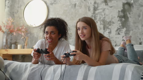 two female friends playing video games lying on the bed. black girl and caucasian young woman holding controllers and having fun.