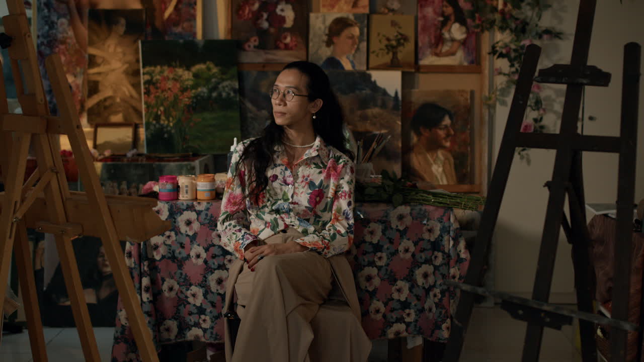 Artist in floral shirt seated in cozy studio with paintings around