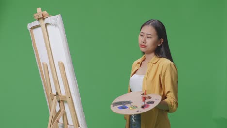 아시아 여성 예술가가가 머리를 흔들고 엄지손가락을 내려놓고 녹색 스크린 스튜디오에서 오일 페인트와 브러쉬로 캔버스에 그림을 그리고 있습니다.