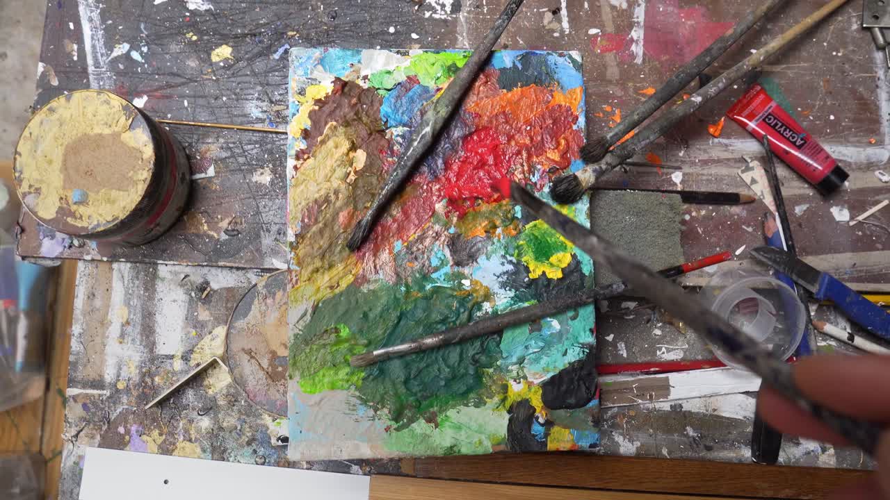 Paintbrush dabbing red paint on color palette top down view on messy table.