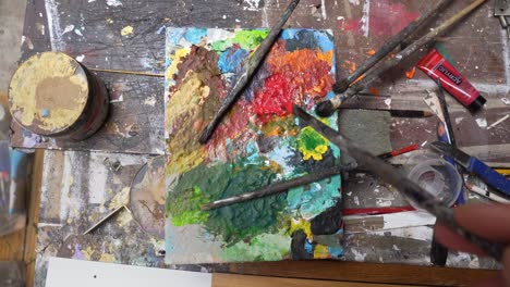 paintbrush dabbing red paint on color palette top down view on messy table.