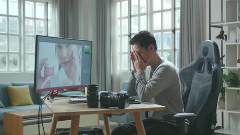 asian cameraman in long sleeved t-shirt and black pants being tired while using desktop computer for editing photo at home.