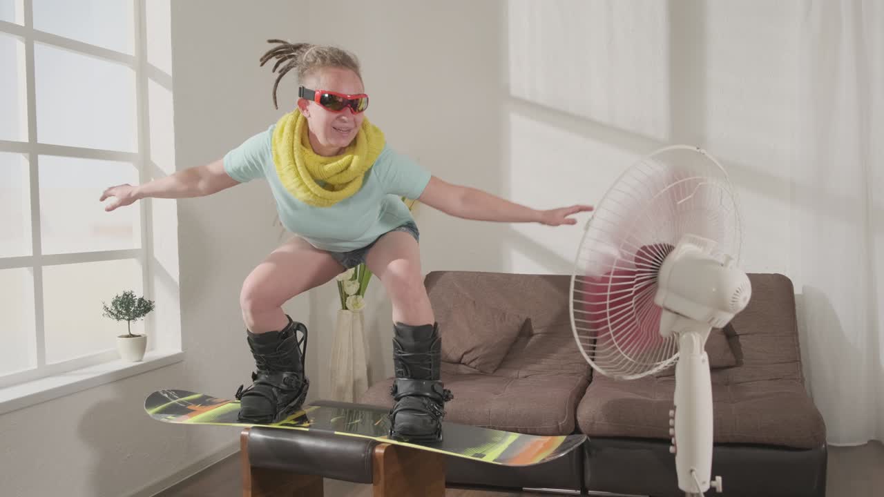 una mujer de snowboard imita el snowboard en la sala de estar de su casa soñando con un viaje a la estación de esquí durante el bloqueo debido al coronavirus.