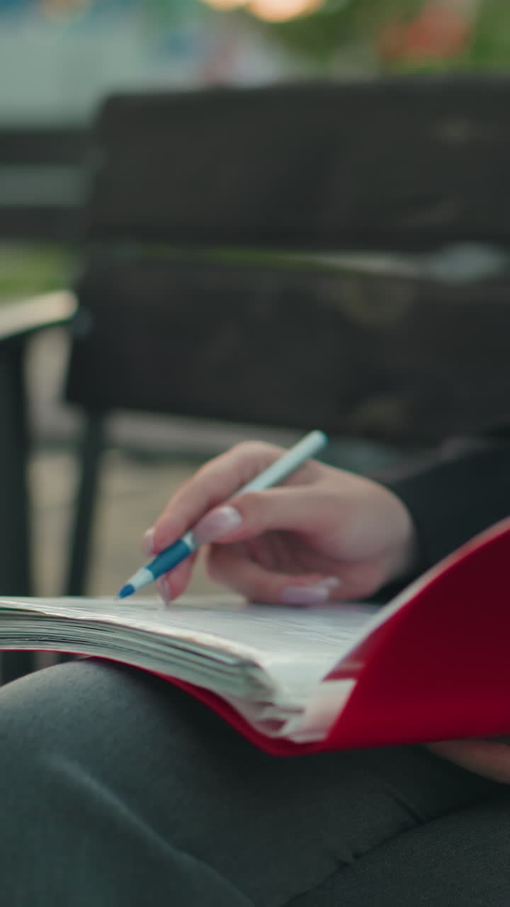 Close up of woman in professional dress reviewing documents in red folder with pen in hand while seated outdoors beside laptop with blurred background featuring benches and greenery