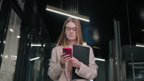 mujer de negocios sonriente mirando el teléfono celular en el interior. mujer de negocios sorprendida leyendo un mensaje en el teléfono móvil en el pasillo de la oficina. mujer mirando la pantalla del teléfono inteligente en el centro de negocios. oficina caminando