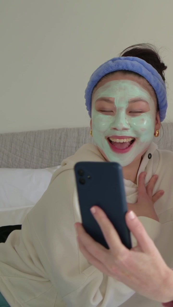 Video verticale: Donna con maschera verde sul letto che si fa dei selfie, mentre un'amica le mostra il telefono, condividendo risate.