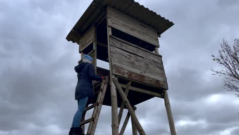 Young-woman-climbing-up-wooden-raised-hide-for-hunting-in-countryside-during-dramatic-cloudy-day-in-wilderness