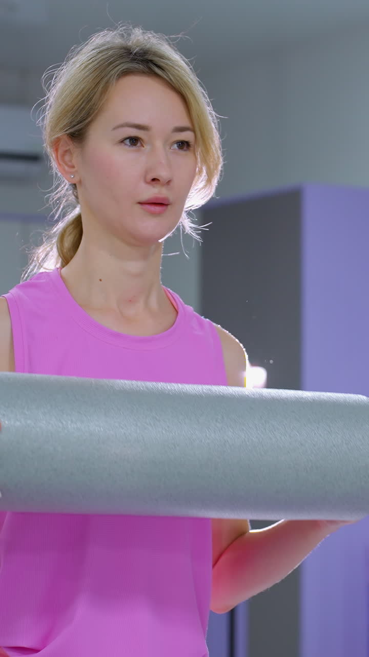 Close-up of a woman stretching forward holding a foam roller, with another woman nearby, bright light and gym equipment are visible in the background