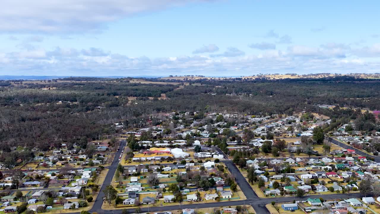 Drone camera glides over suburban Coonabarabran, New South Wales, revealing houses, streets, and surrounding bushland under bright, partly cloudy daylight with smooth forward motion