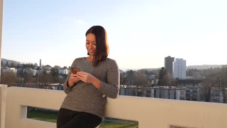 Young-woman-casually-leans-against-the-railing-of-a-balcony-and-smiles-while-typing-on-her-smartphone---American-shot