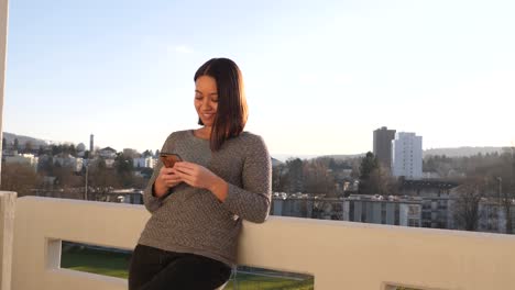 una joven se inclina casualmente contra la barandilla de un balcón y sonríe mientras escribe en su smartphone - tiro americano