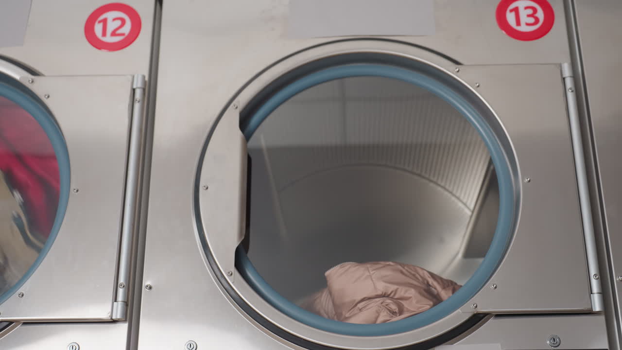 Zoom out view of dryer showing cloth tumbling during dry cycle, drum spinning inside laundromat, moisture evaporating on glass, airflow visible, rotation of fabric through blue gasket ring