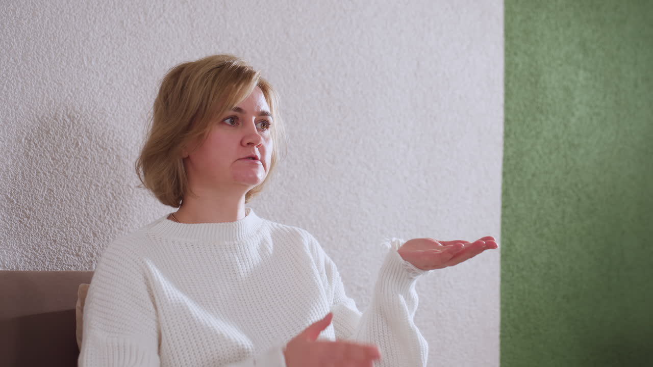 Woman seated on comfortable couch in calming room, demonstrating with her hands while speaking to unseen listener, white sweater and beige pants, cushion behind, green accent wall in background