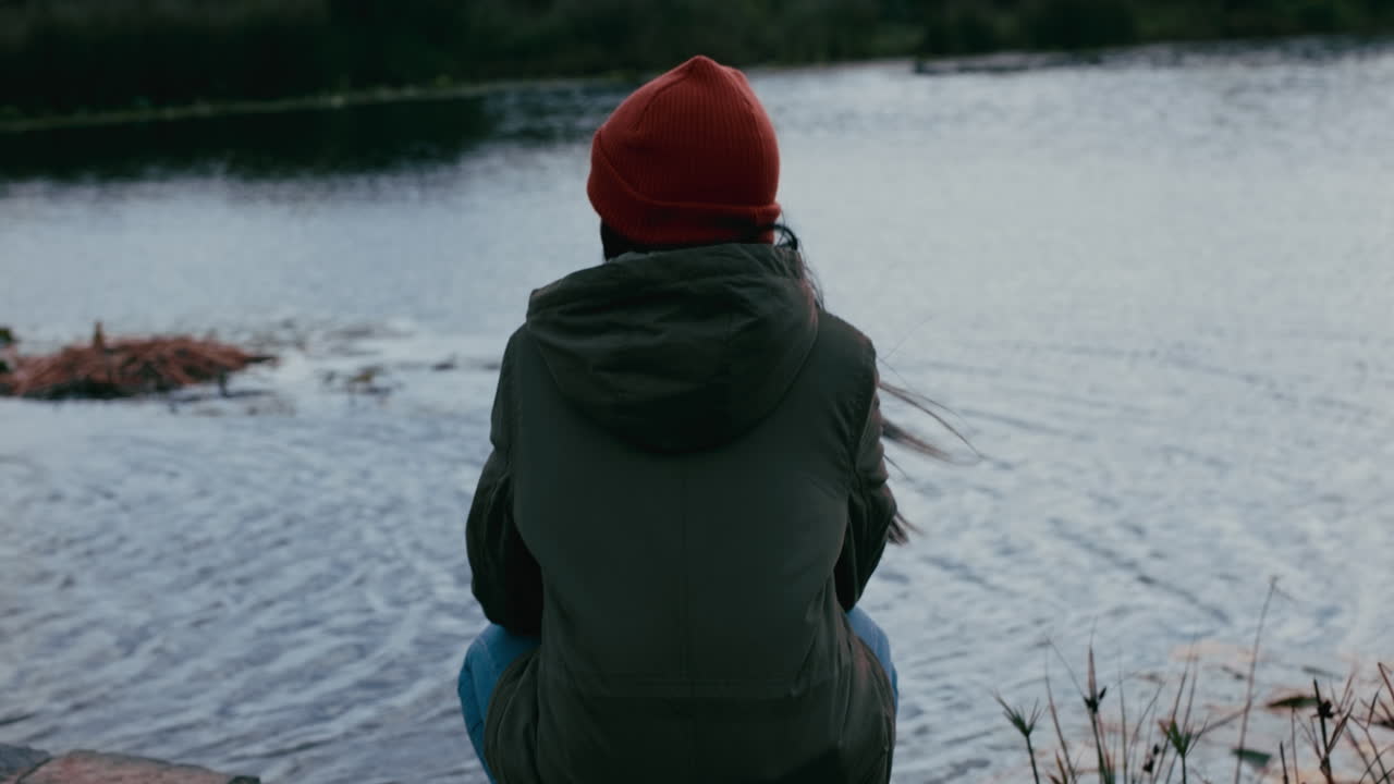 giovane donna solitaria seduta nel parco godendosi un tranquillo laghetto naturale ragazza adolescente che si sente depressa durante una giornata nuvolosa con i capelli mossi dal vento