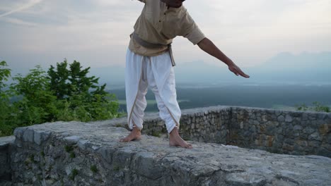 adult indian man rising form a hatha yoga pose doing a stretch at sunrise on castle walls outside in nature in traditional yogi clothes