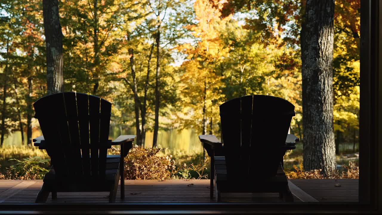Two Adirondack chairs on a wooden deck overlooking a serene forest and lake during autumn