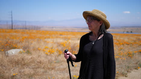 An-older-woman-with-a-walking-stick-hiking-in-a-field-of-flowers-under-a-blue-sky-in-slow-motion