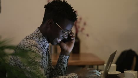 Un-Joven-Sonriente-Hablando-Por-Teléfono-Inteligente-Y-Usando-Una-Computadora-Portátil-En-Un-Café