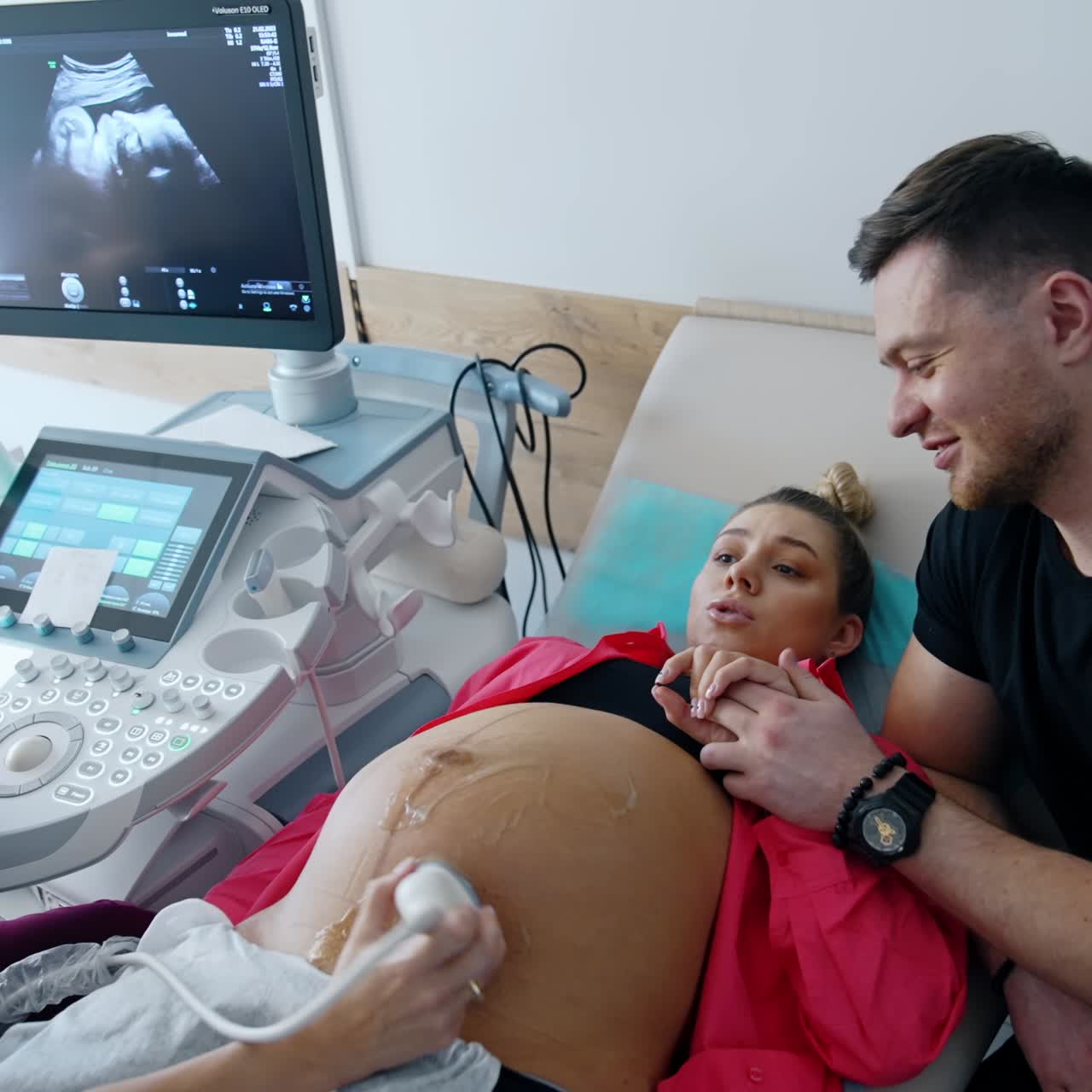 Caucasian expecting parents is in doctor's cabinet. Pregnant woman is checked on ultrasound machine. Smiling man holds his wife's hand
