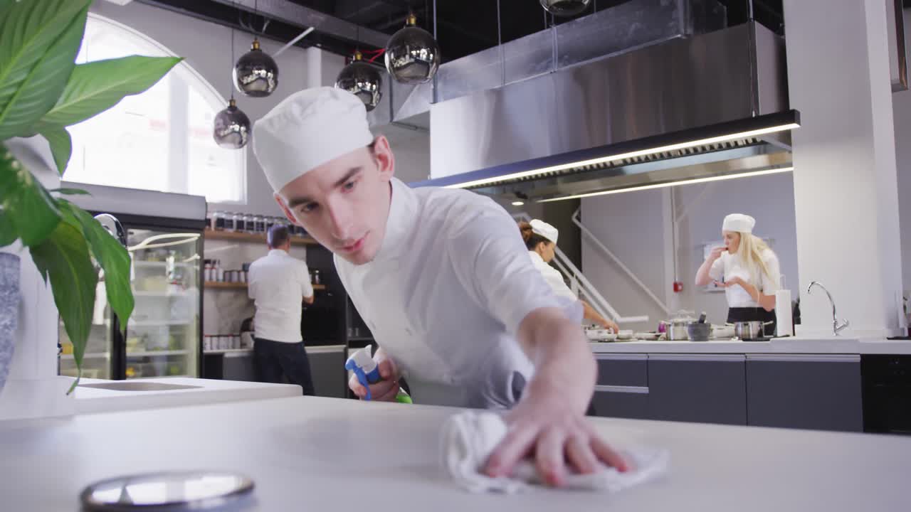 Chef uomo caucasico in una cucina di ristorante che pulisce un piano di lavoro, con colleghi che lavorano